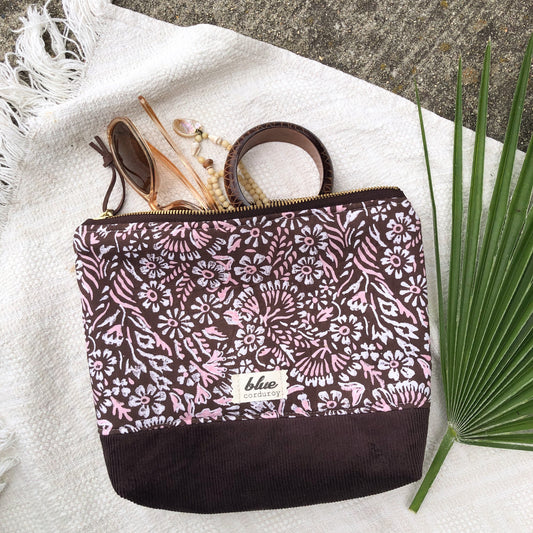 Zipper Travel Pouch made from floral brown fabric and brown corduroy fabric with sunglasses and jewelry coming out and palm branch nearby.
