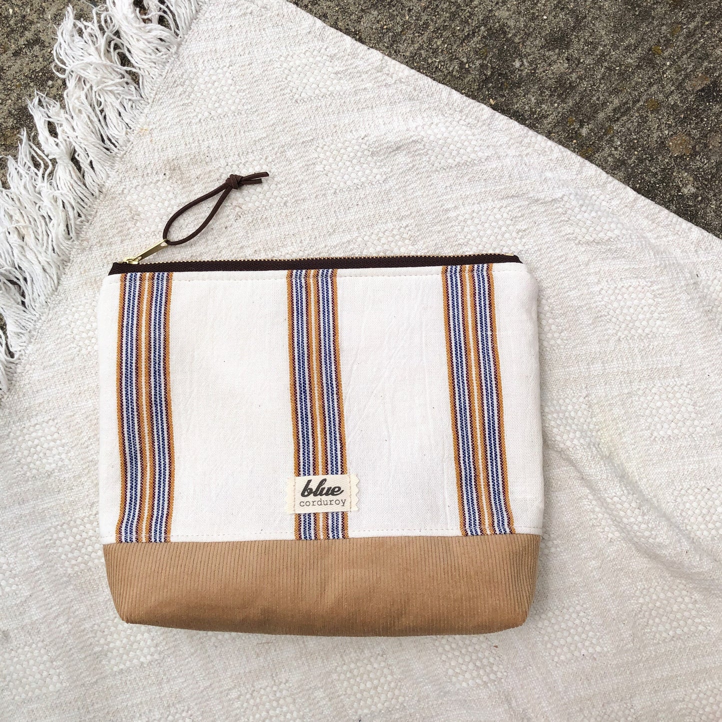 Zipper travel pouch with Blue and tan woven stripes on white background with tan corduroy at bottom laying on white fringe blanket