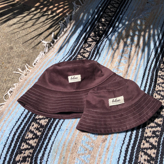 Matching dark brown corduroy bucket hats for adult and baby laying on blue striped beach towel
