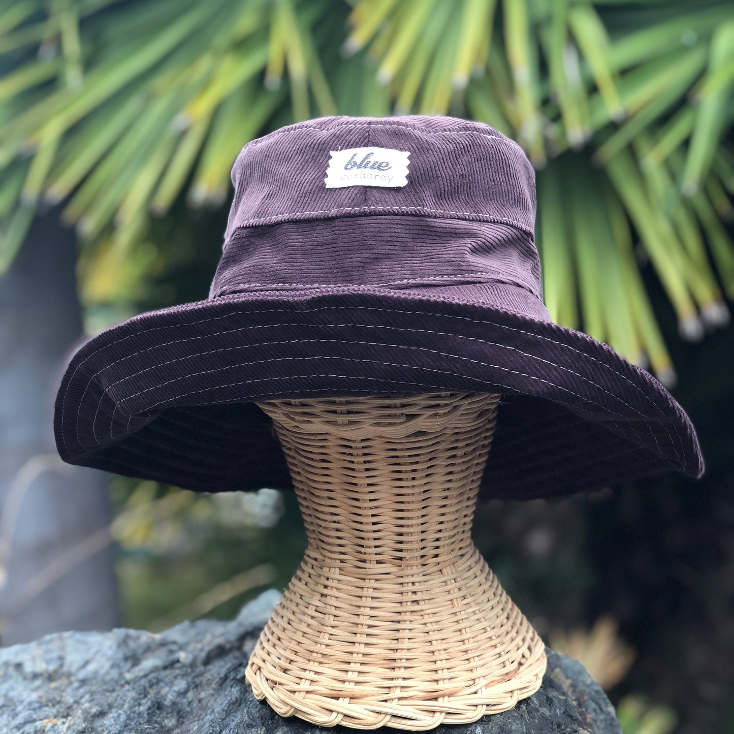 Dark brown wide brim corduroy sun hat on rattan mannequin head outside.