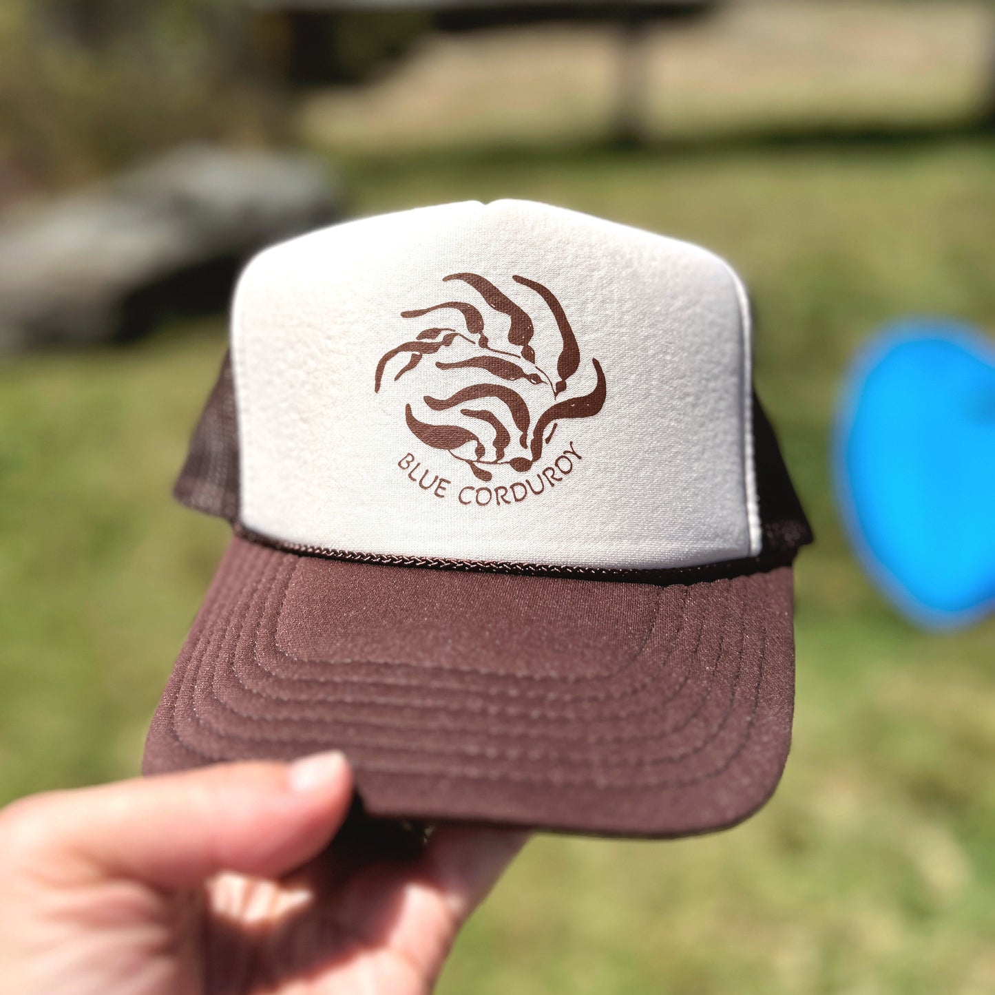 Brown and white cap with 'Blue Corduroy' kelp logo held in front of a blurred natural background
