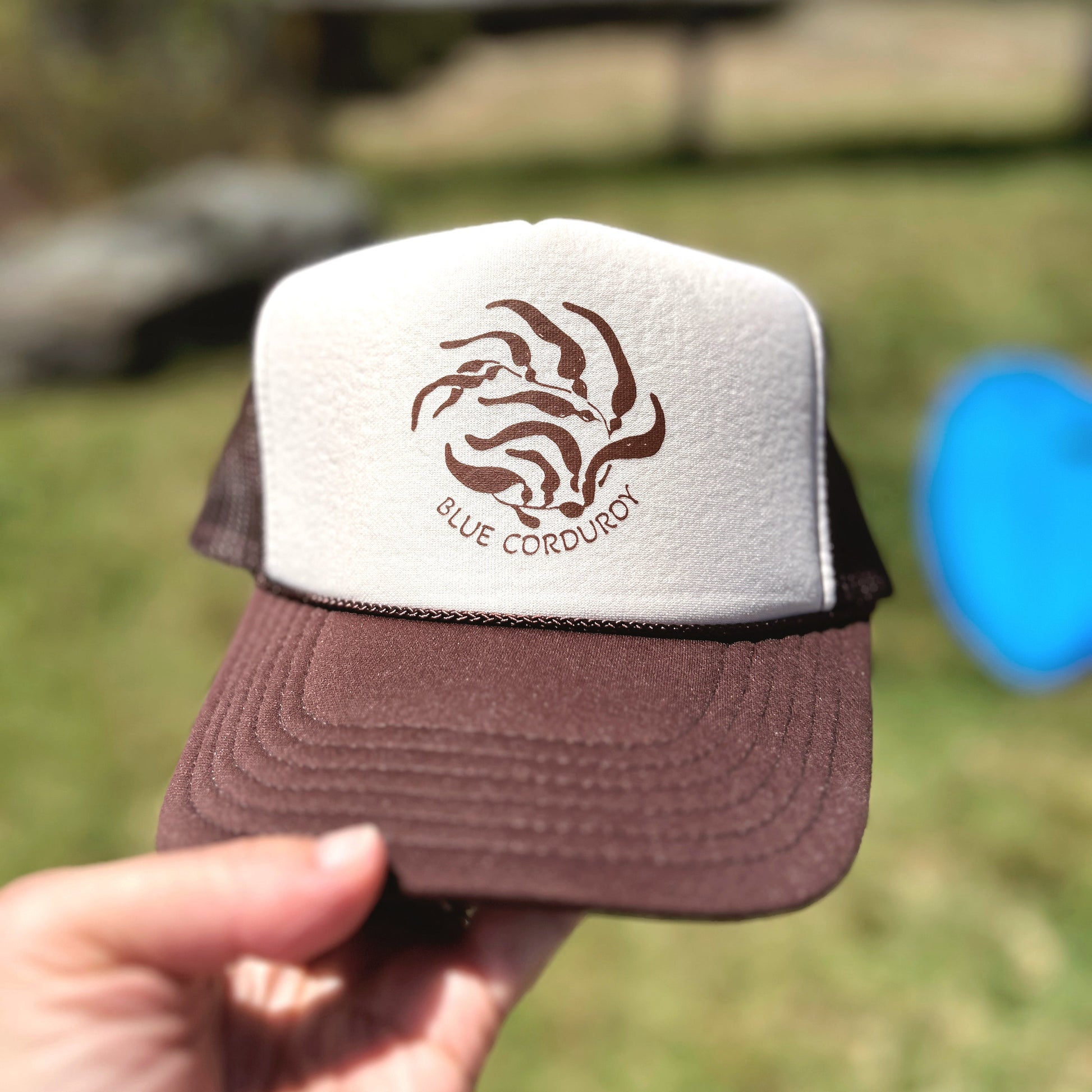 Brown and white cap with 'Blue Corduroy' kelp logo held in front of a blurred natural background