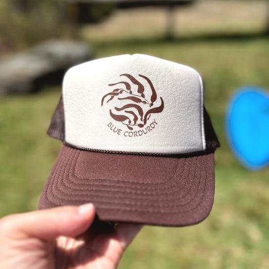 Brown and white cap with 'Blue Corduroy' kelp logo held in front of a blurred natural background