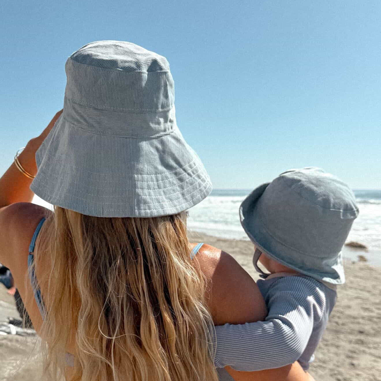 Woman holding a child on a beach wearing matching blue bucket hats.