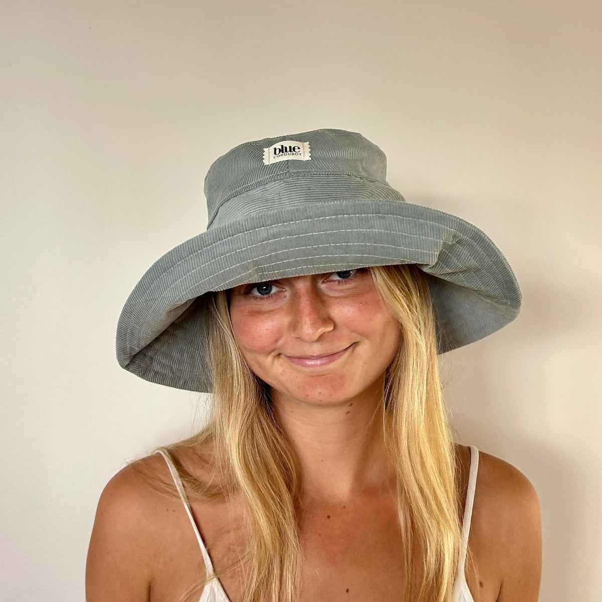 A woman with long blonde hair wearing a wide-brimmed gray sun hat with a small white brand patch. She is wearing a white spaghetti-strap top against a plain, light beige wall.