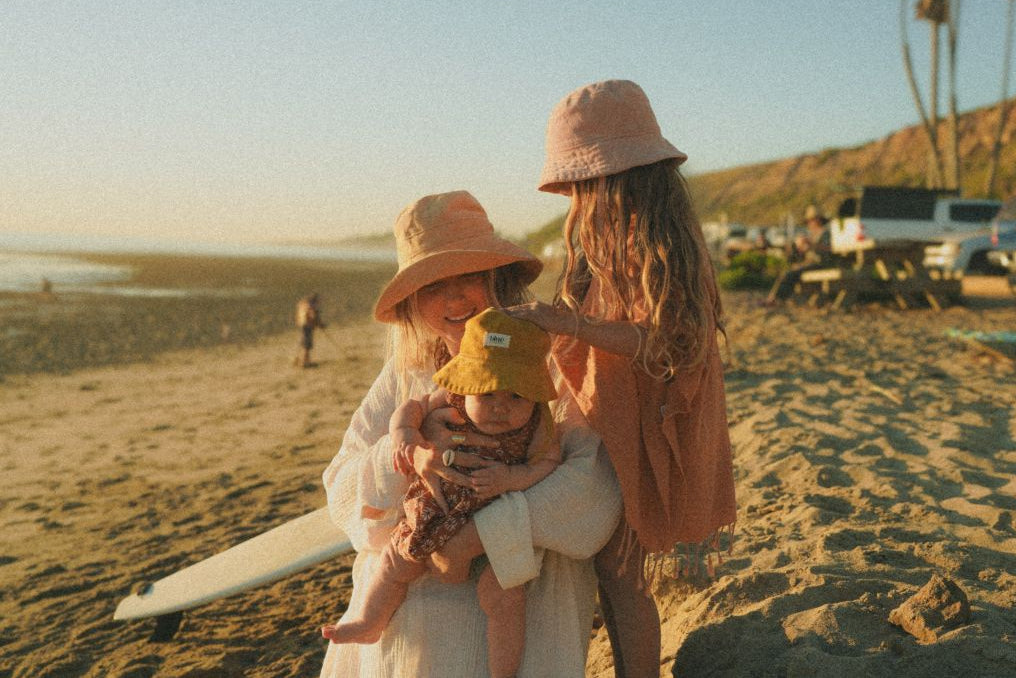 A women and and two children on a beach with surfboards, surrounded by palm trees and vehicles.